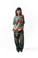 Sollzz शैतान : Inverted Truth Oversize Tee in pastel green featuring a front view of a female model wearing vibrant red and pink abstract graphic artwork prints, styled with printed dark washed jeans and black pointed shoes