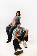 Sollzz शैतान : Inverted Truth Oversize two models posing in matching graphic t-shirts with a green base and red, yellow, and orange abstract swirl patterns, styled with black jeans and sneakers against a plain white studio background.
