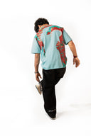 Sollzz Umbra SeaJet Oversize Tee in light blue, back view featuring bold red and teal graphic mask prints, styled with dark washed jeans and a gray baseball cap