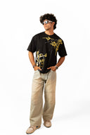 Sollzz Primordia Oversize Tee in black featuring a front view of a male model wearing gold and white abstract face line art graphic prints, styled with beige wide-leg pants, beige sneakers, and white futuristic sunglasses