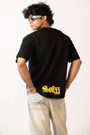 Sollzz Primordia Oversize Tee in black, back view of a male model showing the yellow gothic Sollzz branding print at the bottom hem, styled with beige wide-leg pants and silver sunglasses