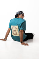 Sollzz - Abyss : Inner Descent Oversize Tee in muted teal, back view of a male model sitting on a studio floor displaying the large cream graphic patch back print, styled with black pants and a patterned bandana